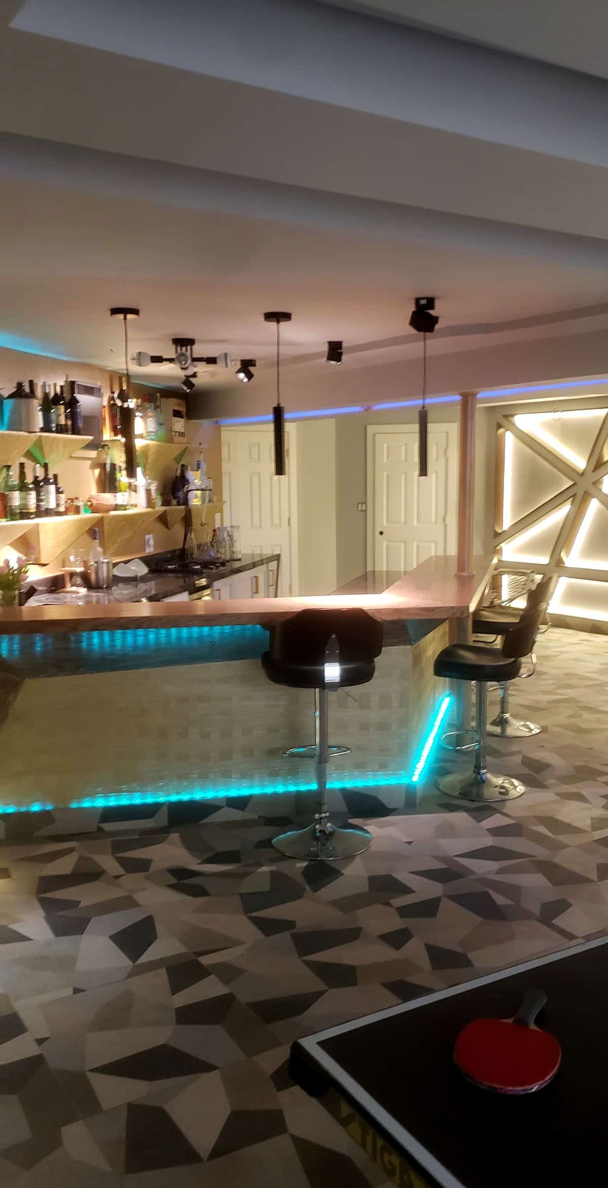 Basement bar counter with stools, cyan LED underbar lighting, gold geometric shelving on left, LED backlit feature panel on right