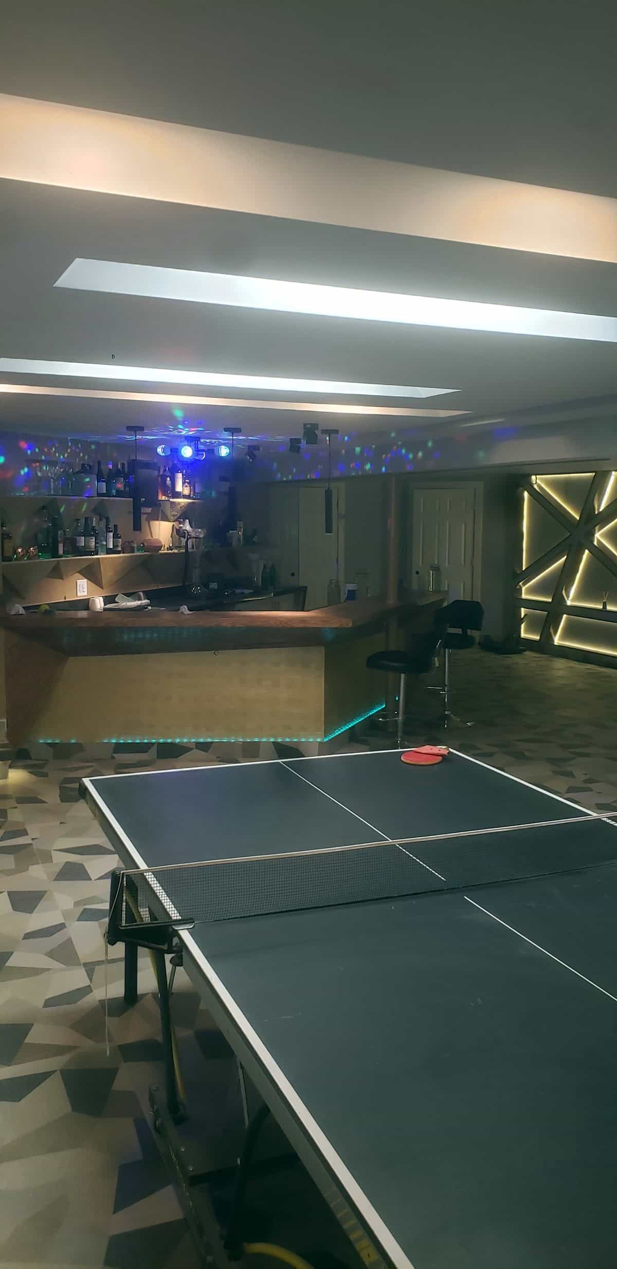 Completed basement entertainment room: ping pong table, bar with cyan LED and active party/disco lights, LED geometric feature wall on right