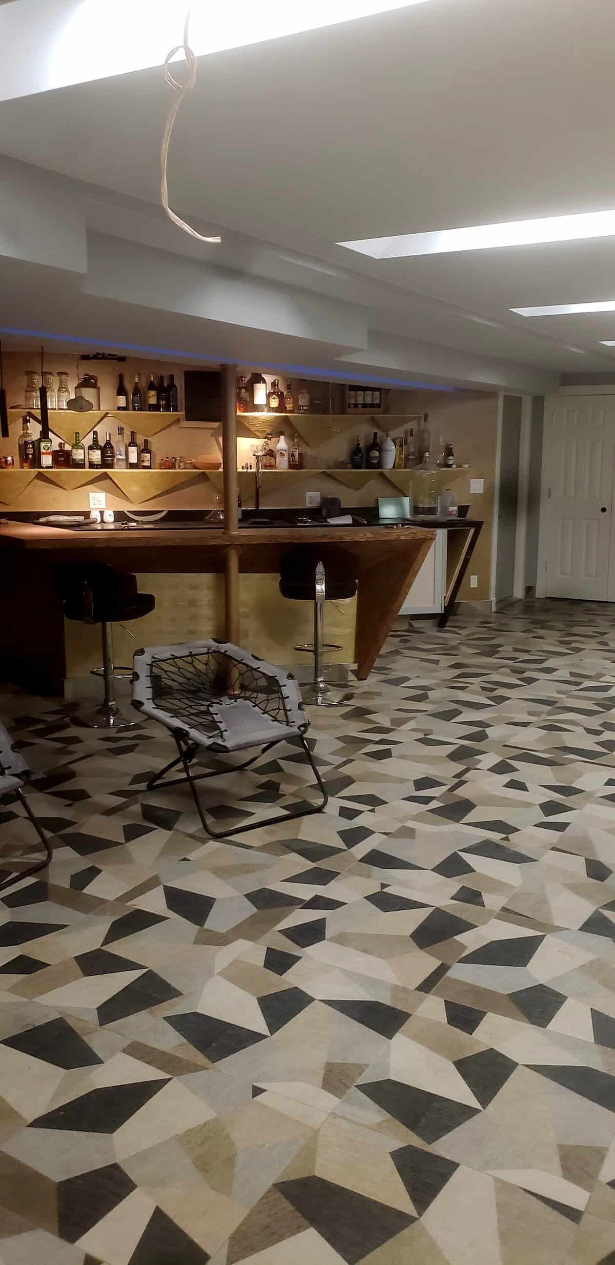 Basement entertainment room wide view: custom gold bar with bar stools, geometric mosaic floor, bungee lounge chairs