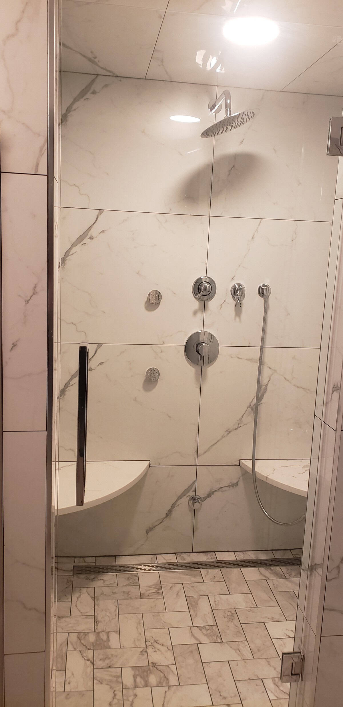 Finished luxury shower with Calacatta marble porcelain walls, chrome rain head and multi-jet fixtures, herringbone marble floor, built-in corner seats