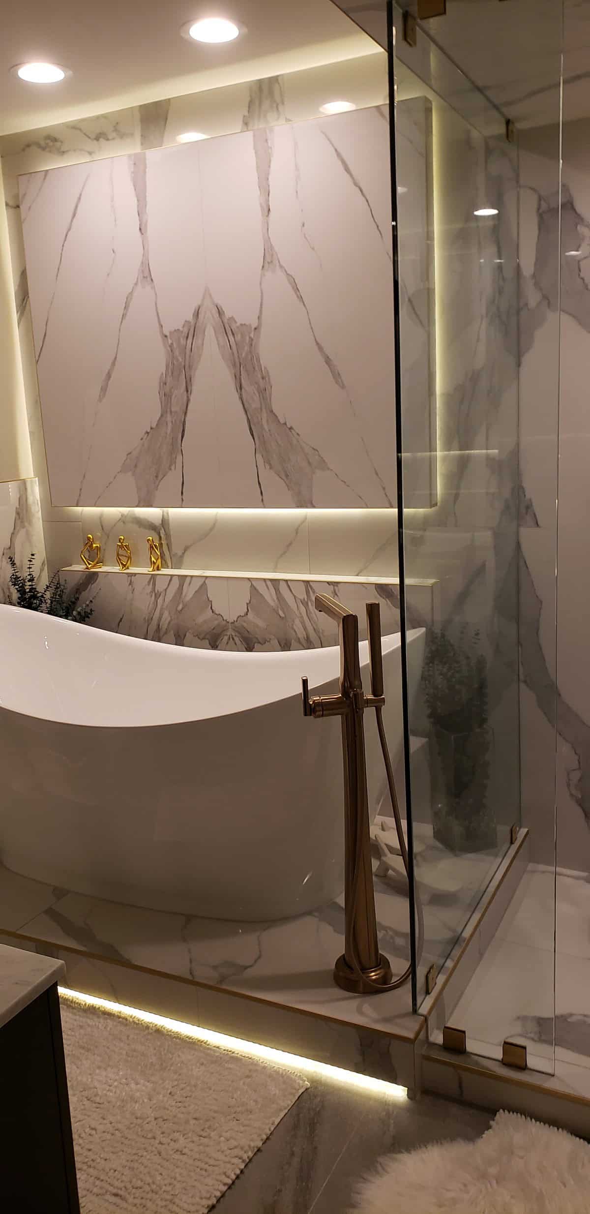 Bookmatched marble feature wall with LED cove lighting, freestanding sculptural tub, brushed gold floor faucet, glass shower, white shag rug, fully finished luxury bath