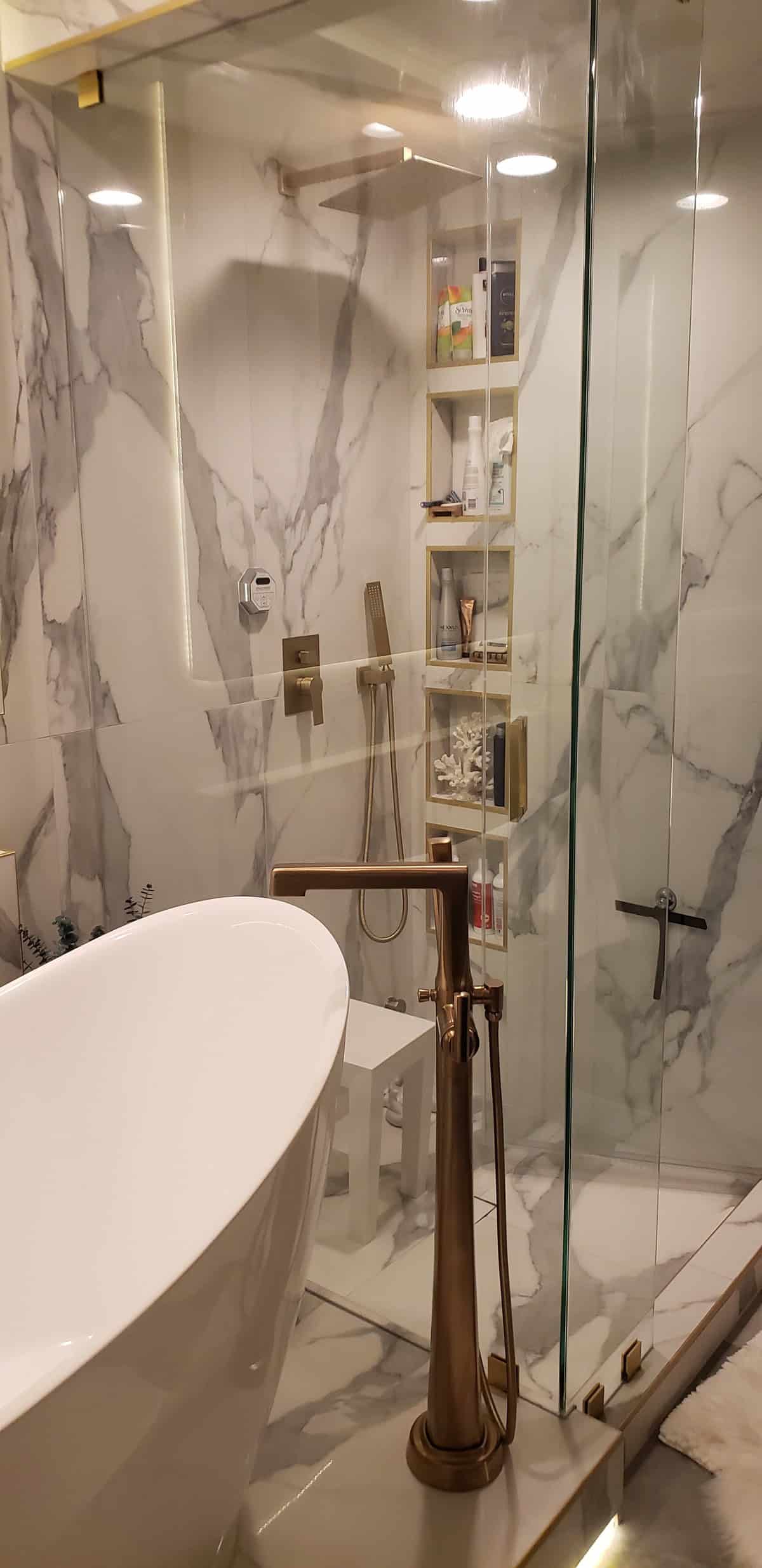 Glass shower interior with bookmatched Calacatta marble walls, stacked gold-trimmed niches, brushed gold floor faucet and tub