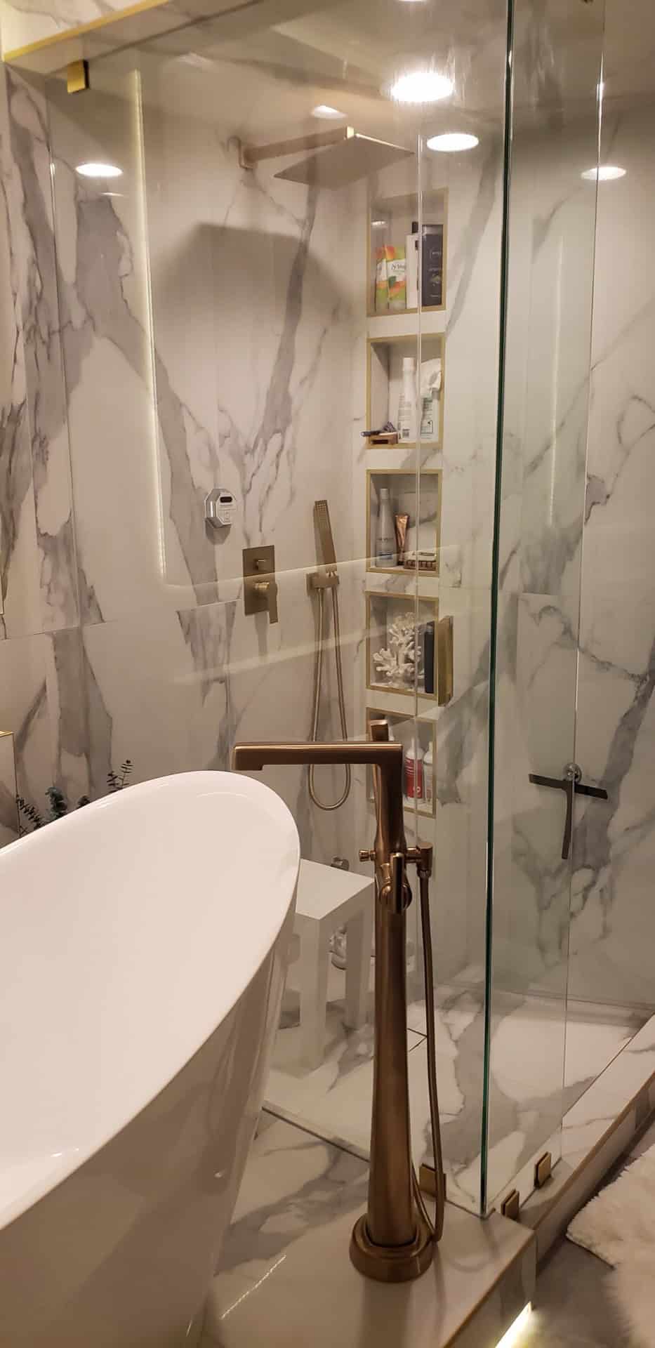 Glass shower interior with bookmatched Calacatta marble walls, stacked gold-trimmed niches with personal items, brushed gold floor faucet and tub