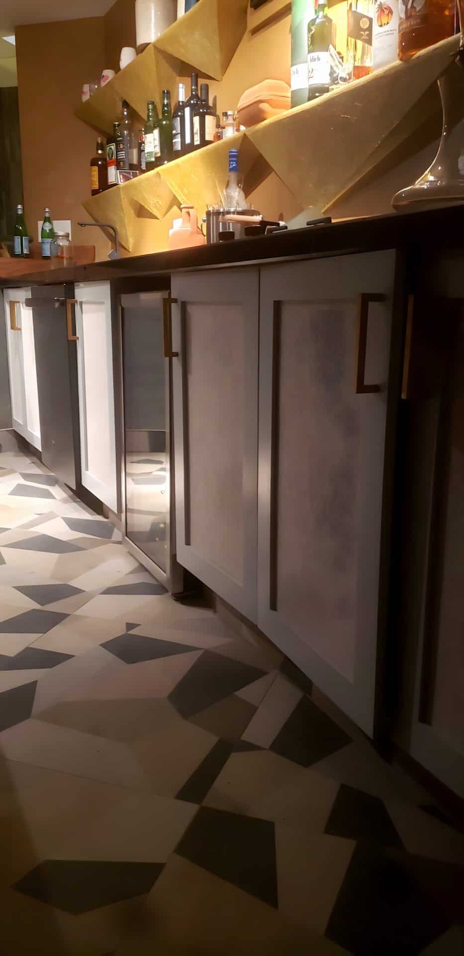 Basement bar side profile: custom white cabinetry with gold bar-pull hardware, wood butcher block bar top, gold geometric shelving with liquor bottles