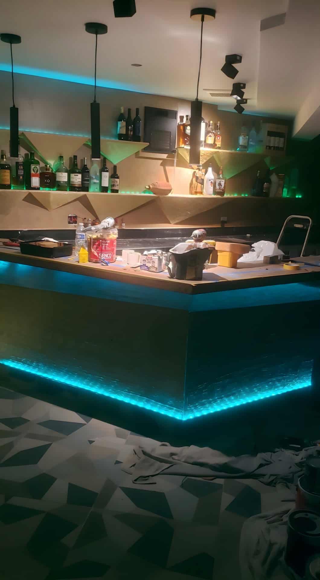 Basement bar with cyan LED undercounter lighting, green LED geometric gold shelving, pendant lights, liquor bottles