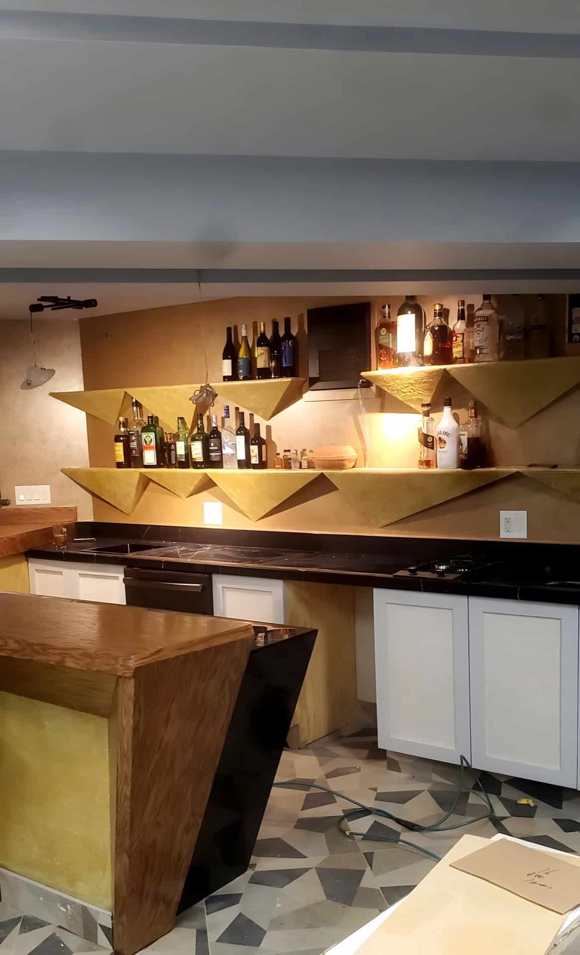 Basement bar wide shot: custom gold geometric angular shelving with liquor bottles, black granite bar top, white lower cabinets, geometric mosaic floor