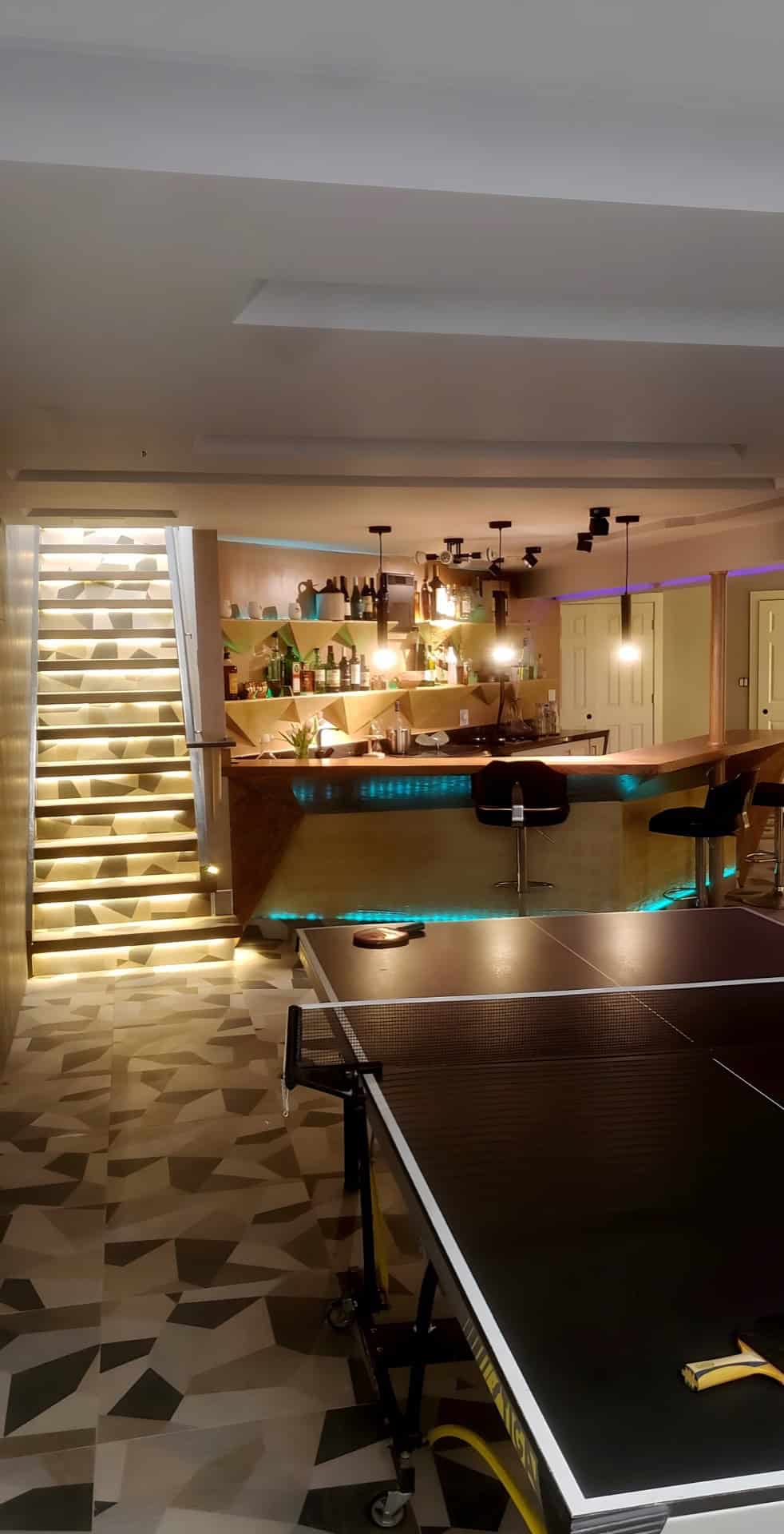 Staircase with LED-lit mosaic base beside a basement bar with gold shelving — custom staircase installation