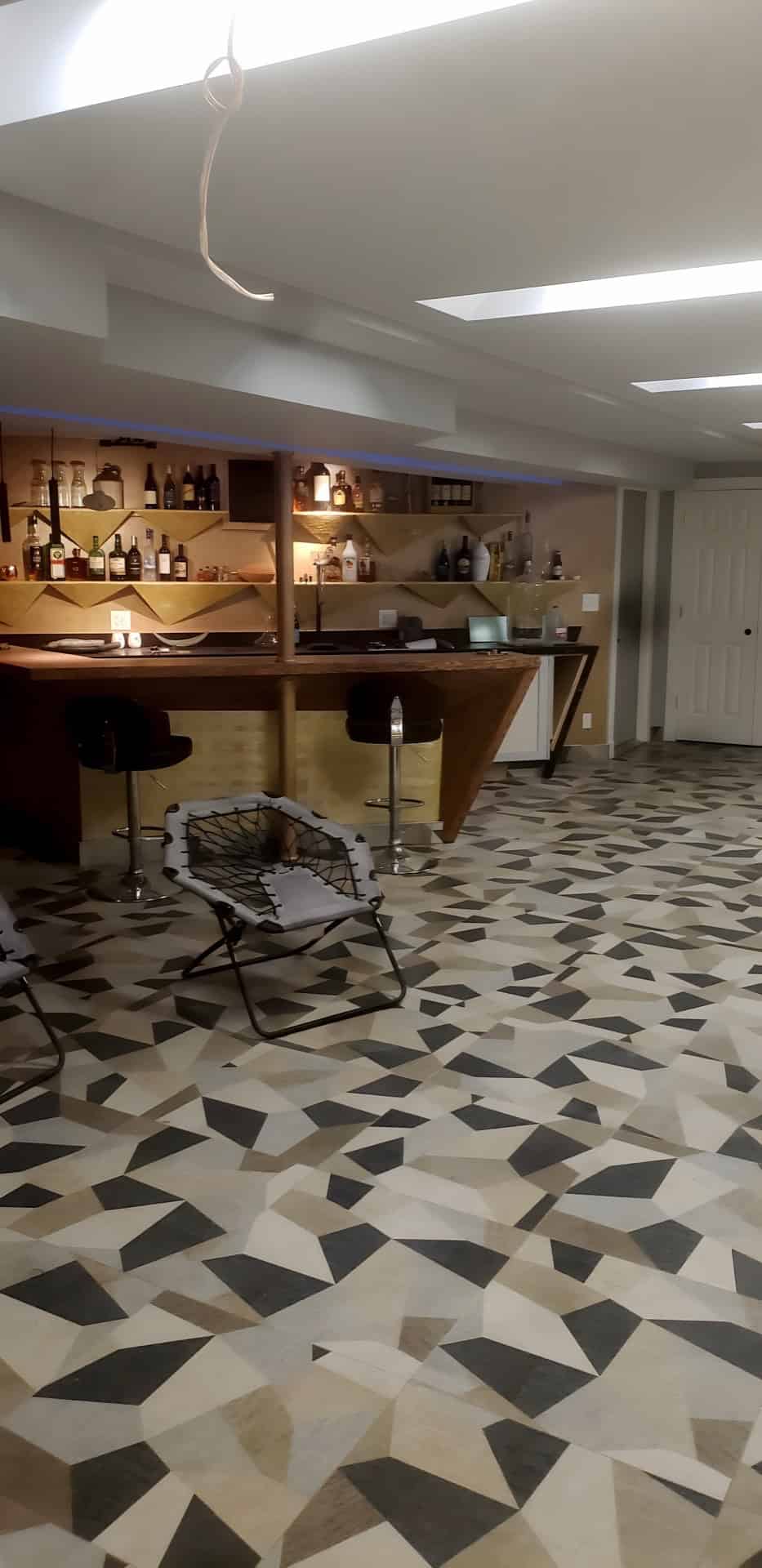 Basement entertainment room wide view: custom gold bar with bar stools, geometric mosaic floor, bungee lounge chairs