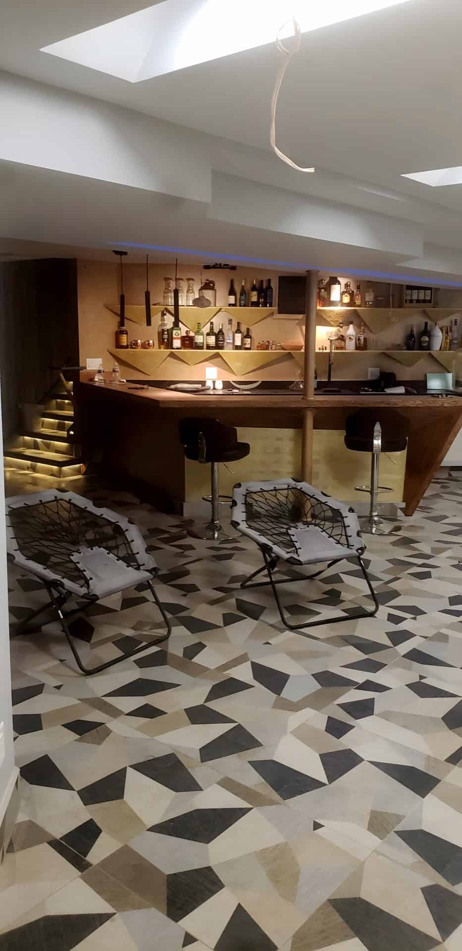 Basement bar area with LED-lit mosaic staircase visible in left background, gold geometric bar and bungee chairs