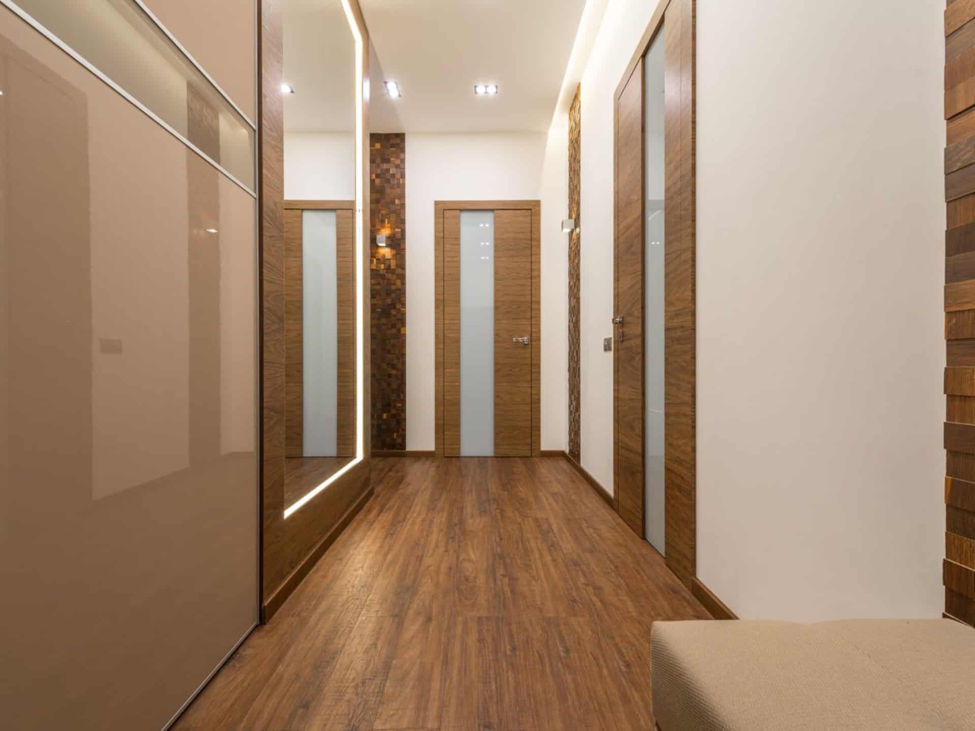 Hallway lined with wood-veneer interior doors with frosted glass inserts and mirrored wardrobe doors over plank flooring